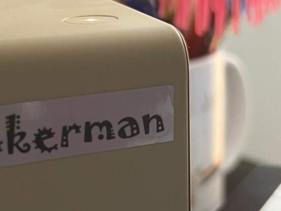 A photo of a small label stuck to the front of the label maker. The text “kerman” is visible in a funky font. The top right corner is a little dog-eared and not stuck down.