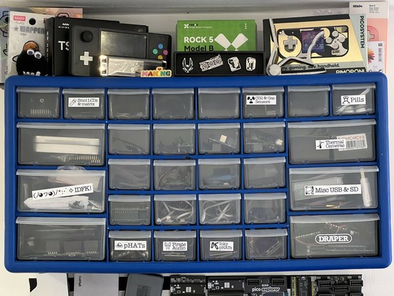 A photo of a blue component draw unit with clear drawers. There are 24 small and 6 large drawers of which only 9 are labelled with various styles of black on white label. On top of the drawer is a clutter of small boxes and curios.