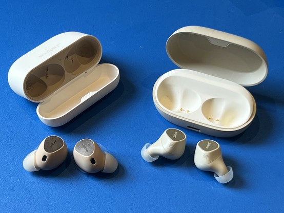Two sets of earbuds. On the left are the Elite 10 in front of their taller, thinner case, with flattened ear
tips and in a dark beige. On the right are the Elite 7 Pro with starkly elongated ear tips, in a lighter beige and with a wider, flatter box. Both are sat on a bright blue table.