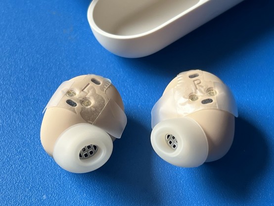 Closeup of both of the Jabra Elite 10 earbuds sat button-down on a blue table. There are clear L and R labels embossed on the buds, and a paper wrap covers these and the charging contacts for shipping. The ear tips are oval-shaped and have a coarse silicone grid to catch debris. The buds are dark beige.