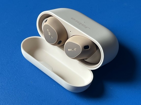 A plastic charging case laid down on a blue table with the lid open, revealing two dark beige earbuds nestled in their charging cradles. The case is flat and tall, with a rounded bottom that prevents it standing upright. A single pin-prick LED sits between the earbuds and another on the outside of the case.