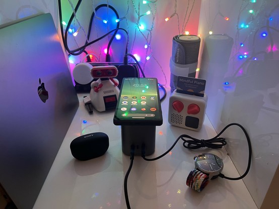 A Sony Xperia 5 V charging wirelessly on top of UGREEN’s Nexode 2-in-1 desktop charger. It’s sat approximately in the middle of a gloss white cubby/shelf surrounded by an assortment of gadgets. There’s a 1980s Tony OMS-B Dingbot (a tiny red and white robot toy) in the background for some reason.