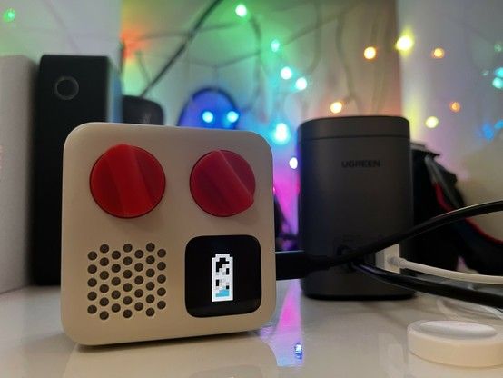 A tiny white audio player with two orange dials, a speaker grille and an LCD screen plugged into UGREEN's desktop charger via a USB cable.