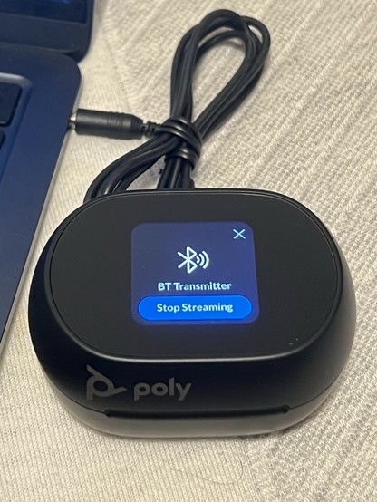 The Poly Free 60+ UC charging case. It’s a little black oval shaped thingy with a tiny colour LCD screen on top showing “BT Transmitter” and a button reading “Stop Streaming.” An audio cable, still tied in a neat bundle, connects the charging case to a laptop via the 3.5mm audio Jack.