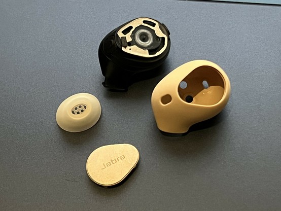 A single black earbud sat next to its little oval ear tip, it’s button and it’s silicone rubber outer shell. The shell looks a little like a startled face. underneath the removed button cap the actual button is revealed, a tiny, round dome switch that looks like it could be water sealed.