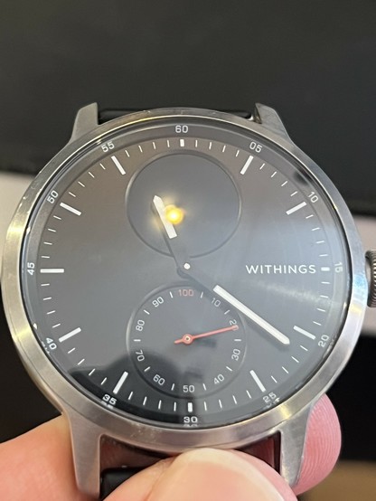 A photo of ScanWatch 1. It depicts a steel watch body with a chamfered edge leading to a glass cover with a black face behind it. The face is marked with numbers at five minute intervals and has white marks for hours and minutes. There are slender, pointy, hour and minute hands but no second hand. Underneath the hands are two complications- the upper is an OLED screen (currently off) and below that is a secondary dial with numeric markings at intervals of 10, up to 100, to indicate step goal progress. The 100 and the dial hand are picked out in contrasting red.