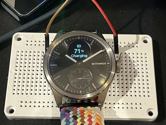 A photo of a steel and black watch with a rainbow woven strap. It’s sat atop a white electronics breadboard with a brown and red wire sticking out and disappearing out of the shot. Red is on the left (the watch body) and brown on the right (the crown). Two little strip headers are plugged into the breadboard to act as spring contacts to make a connection with the watch body and crown. The little OLED display says “71% Charging.”