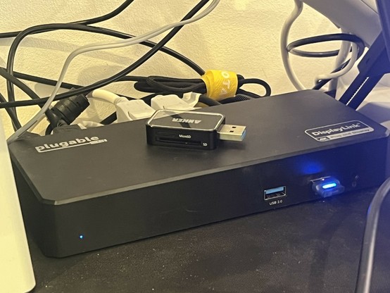 A black USB Type-C docking station with a nest of wires erupting from the back. In the front. There’s some kind of wireless dongle, lit up blue. On the top there’s a microSD card reader. A logo in the top left reads “plugable” and the bottom right “DisplayLink.”