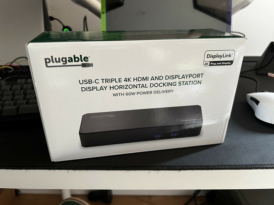 A white box with a picture of a wide, flat, black, USB Type-C docking station on top. A title reads “USB-C Triple 4K HDMI and DisplayPort Display Horizontal Docking Station” with smaller text below reading “With 60W Power Delivery.” Yes,
It really is that verbose. A “plugable” logo adorns the top left of the box and “DisplayLink” the top right.