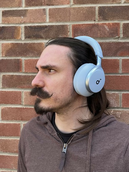 A photo of me, a moustachioed weirdo with a goatee and long hair wearing a reddish brown hoodie and stood in front of a brick wall. The sea of brownish reddish tones are starkly offset by a pair of bright blue headphones adorning my head. They’re chunky, and have a little “d” shaped logo - The “d” from SoundCore - which also hints at musical notation.