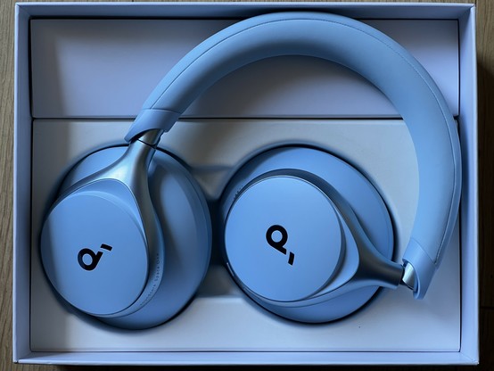 A photo of a pair of baby blue headphones folded up in their box. They are sat such that both the outer sides of the large, over-ear cups face upwards. There’s a little “d” logo on each, stylised to look perhaps a little like musical notation. The matt baby blue plastic and faux leather is offset by a semigloss, metallic-blue finish where the cups join to the headband.