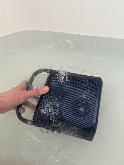 A small, dark blue portable Bluetooth speaker half submerged in a bathtub. It’s playing incredibly bassy music causing little jets of water droplets to erupt about an inch or so from the speakers. The “d” logo of Soundcore is clearly visible on the front.