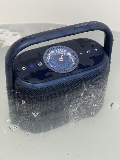 A tall, blue, Bluetooth speaker submerged in foamy bath water. The top and handle jut out above the water, but the top speaker has very obviously got a pool of water in it. The lights are illuminated showing it’s still working. Diffraction makes it look much squatter and smaller than it actually is.