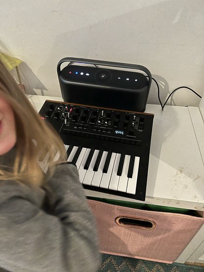 A small electronic synthesiser sat atop a white kallax shelving unit. Behind it sits a wide squat, curvy speaker connected via a 3.5mm audio cable. There’s a motion blurred child just out of shot in the foreground.