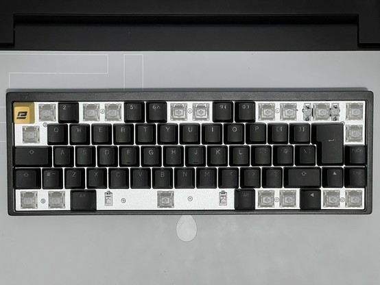 A top down photo of the KB65HE keyboard showing 18 key caps removed to reveal the retaining screws underneath. Beneath the caps is a bright aluminium plate. There are ten screws.