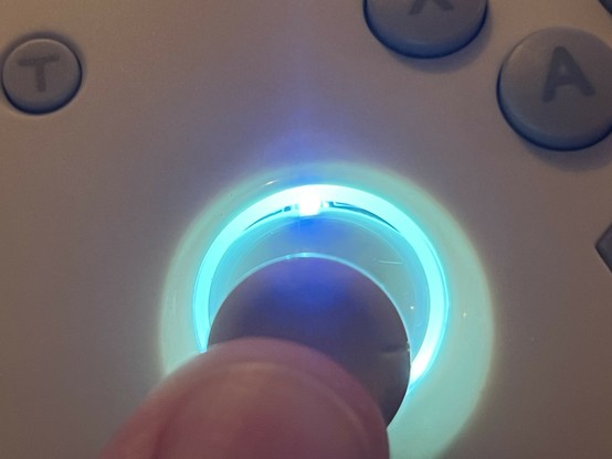 A closeup of the thumb stick being tilted down by a thumb. The resulting gap reveals the LED beneath, lit up a lovely turquoise but glaring brightly with no diffusion.