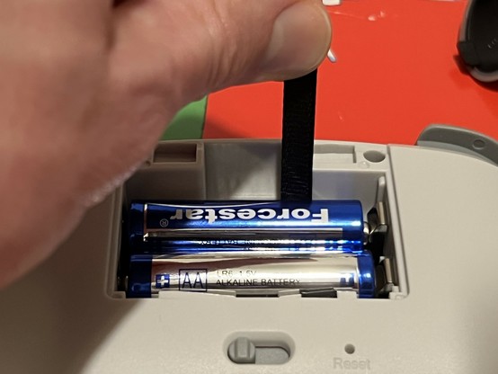 A photo of the battery compartment in a cheap GioTeck controller. Some fingers are grasping and pulling the little fabric battery release pull.