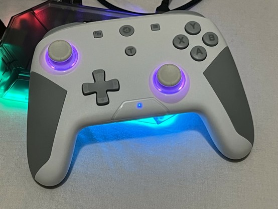 A white gamepad with medium grey buttons and rubberised portions of the hand grips. The analog sticks - in an Xbox style configuration - are backlit purple. The light is shining through the white plastic too. The D-Pad is cross shaped, but looks more Maltese than Nintendo.