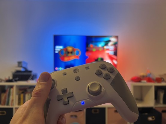 A photo of a white and grey gamepad being held by one hand with a TV blurred out in the background.