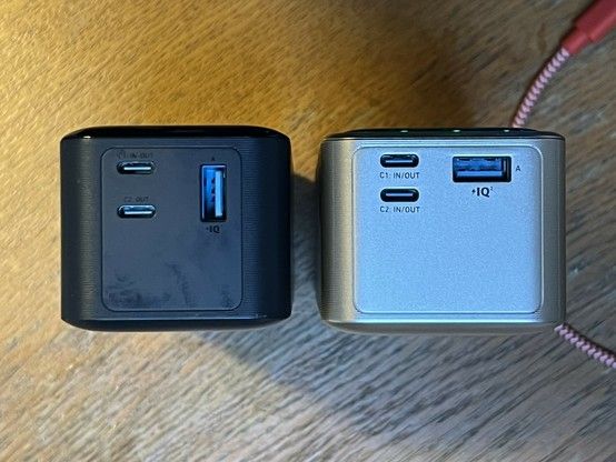 Two portable power banks side by side on a wooden surface, one black and one light gold, each with two USB Type-C ports and one USB Type-A port visible. The newer, gold power bank tucks the USB Type-C ports closer to each other perhaps to help accommodate its seventh battery cell.