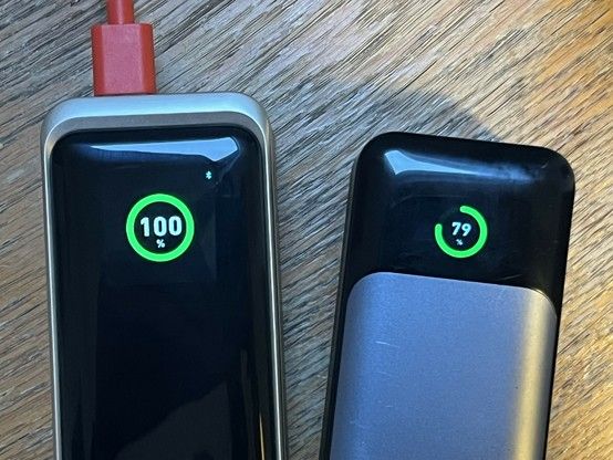Two portable power banks on a wooden table showing battery levels; the left one displays 100% charge with a cable attached, and the right one shows 79% charge.