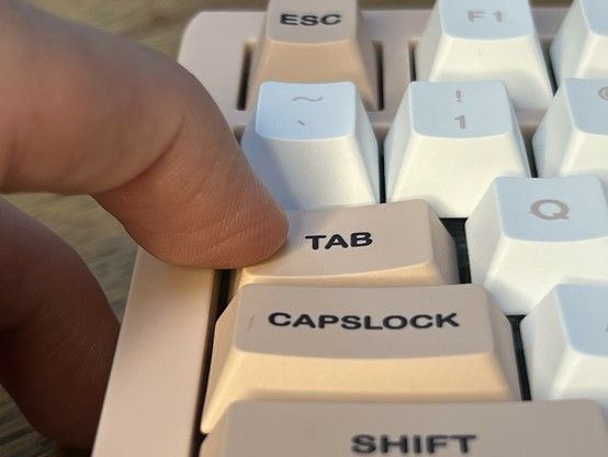 Close up of a mechanical keyboard with white and tan keys. A finger is pressing the Tab key on the extreme left edge, showing that it tilts a little but not as much.