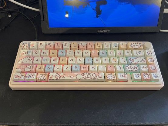 A mechanical keyboard with custom keycaps featuring cute animal illustrations and pastel colors, placed on a black desk mat with a portable monitor in the background. A rainbow backlight leaks out from between the keys.