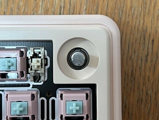 Close-up of a mechanical keyboard's interior showing pink and teal switches and stabilisers protruding from a black PCB with white silkscreen. On the top right is the metal post of a rotary encoder, its cap removed.
