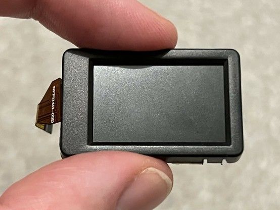A person's hand holding a tiny LCD module, with the flat flex cable hanging out of the left.