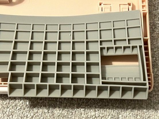 A thick matt-like silicone rubber insert laying unturned in the bottom shell of a pink keyboard. It has a grid pattern of supports that- with the help of harsh lighting from the side- reveal it to be tapered toward one side.