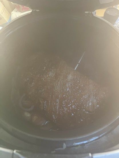 A beef brisket cooking in a rice cooker on slow cook mode. The view is mostly obscured by steam. A silver skewer-like wireless meat thermometer is visible through the haze.