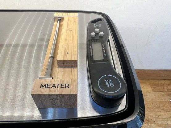 A wireless meat thermometer in a wooden charging block sat next a black, flip out digital meat thermometer on a metallic surface.