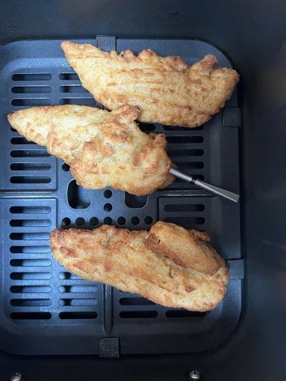 Three pieces of battered chicken on a black grill tray. A wireless meat thermometer- a silver skewer like thing- protrudes from the middle piece.