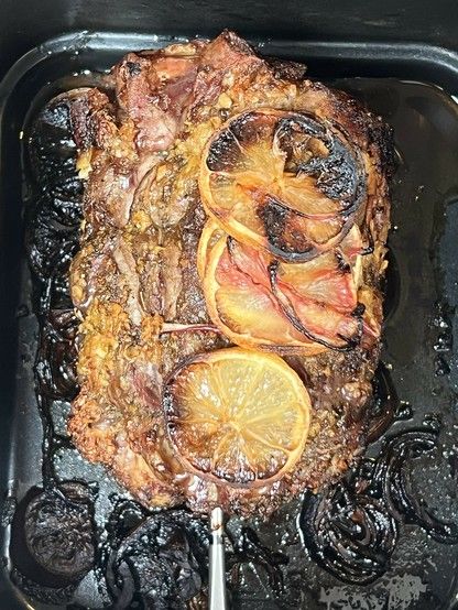Roasted meat with caramelized lemon slices on top, in a baking tray with charred onions. From the side of the meat protrudes a skewer-like meat thermometer.