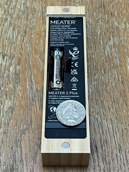 A Meater 2 Plus wireless meat thermometer wooden charging block, with its battery compartment open showing a single AAA battery. On top is a ten pence coin ostensibly for scale but actually covering the serial number.