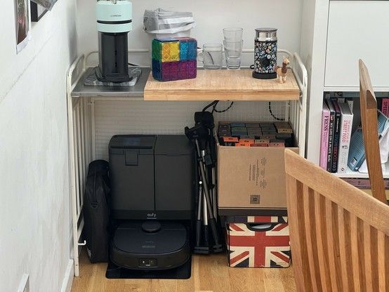 A corner of a room with various household items including a coffee machine on a metal table. Beneath it is a robot vacuum and base station, a storage box with a box of coffee pods on top, various tripods and phone holders, and a ring light case wedged against the wall. There's also a wooden chair in the foreground. The wall is noticeably discoloured by some kind of spill.
