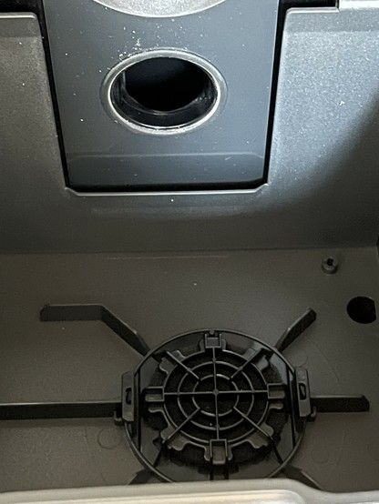 Close-up view of a black plastic fan guard on a piece of electronic equipment, with a hole above it where dust and dirt would be drawn in. Dust particles are noticeable on the surfaces. It’s the inside of a vacuum dust compartment in a RoboVac base station.