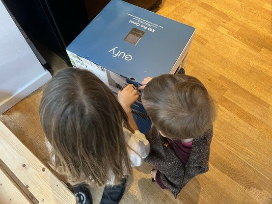 Two children from an overhead perspective looking at a closed cardboard box on a wooden floor. On the box is written “Eufy X10 Pro Omni.”