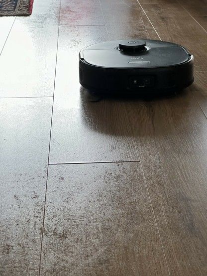 A robotic vacuum cleaner on a dusty wooden floor with a patterned rug in the background. There are noticeable water marks on the floor where it has been mopped.