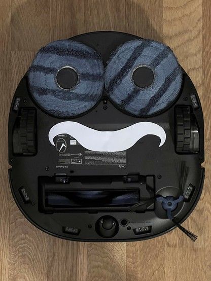 An upside-down robotic vacuum cleaner on a wooden floor. The arrangement of mop pads and vacuum aperture give the impression of a face. A white paper moustache has been cut out and laid over the serial number.