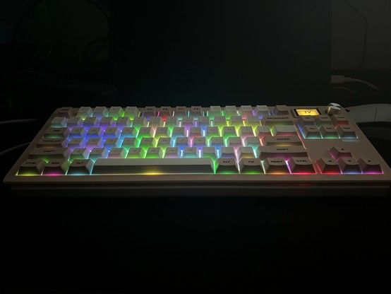 Illuminated mechanical keyboard with multicolored backlit keys on a dark background. A patchwork of pastel coloured lights shines through the cake between the keys, which are picked out with black and white text that is not illuminated.
