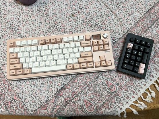 A mechanical keyboard with pink and white keycaps on the left and a separate numerical keypad on the right, both placed on an intricately patterned tablecloth.