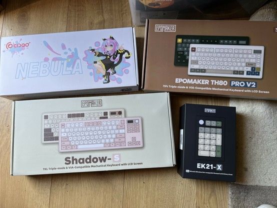 Four product boxes for mechanical keyboards sat on a wooden floor. From upper left to bottom right: “CIDOO Nebula” with anime catgirl art, “Epomaker TH80 Pro V2” with the box art showing black and vintage white variations, “Epomaker Shadow S” with the box showing brown and pink variations and “Epomaker EK21-X” depicting a numpad with green white and grey keys. (Though it’s actually black and pink!)