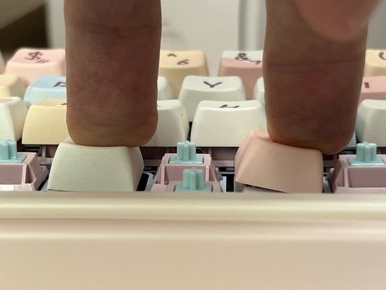 Two fingers pressing the edges of keys on a pastel-colored mechanical keyboard, illustrating the stability of two different switches. The right-most key- using the cheaper “Carda” - is leaning quite far to the right. The left-most - using a “Sea Salt” switch - has much less lean.