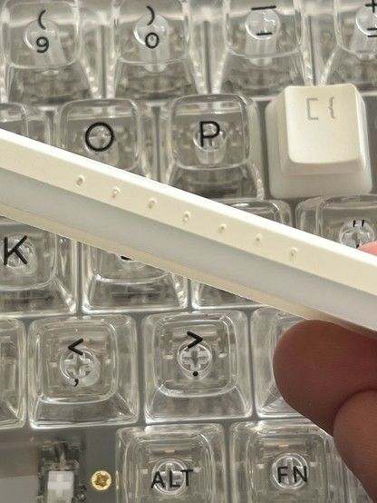 A close-up view of a mechanical keyboard with transparent key switches and one keycap removed, showing the switch underneath. A hand is holding a white space bar in the foreground and showing some nasty injection moulding artefacts in the plastic.