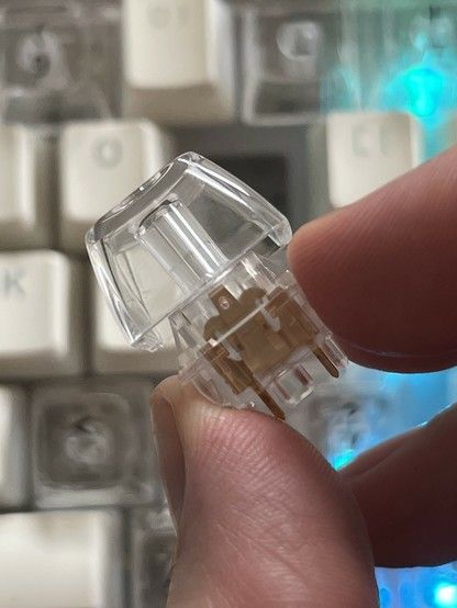 A close-up of a clear mechanical keyboard switch and key held between fingers. The copper switch contacts can be seen inside. White keyboard keys are blurred in the background.
