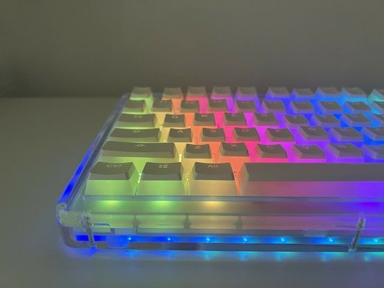 Backlit mechanical keyboard with transparent edges lit up with rainbow light. The keys are translucent frosty white with a white cap on top- resembling Japanese “Purin” custard pudding. The sides of the keys have a pleasing diffuse glow and the legends are also clearly lit up, except with blue light when they’re a little unclear.