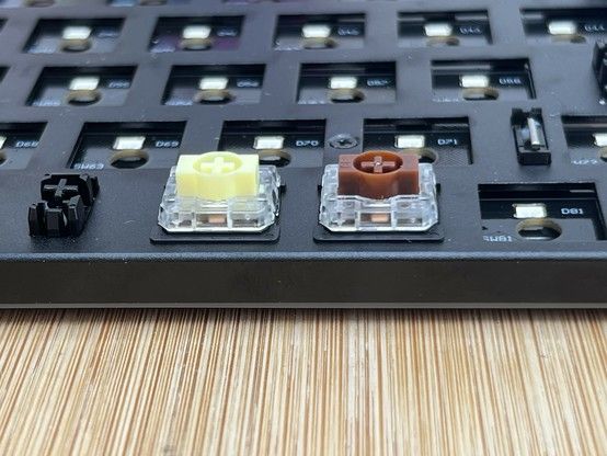 Close-up of a mechanical keyboard without keycaps, showing different types of switches: a black stabiliser is visible on the left, a yellow switch in the middle, then a brown switch on the right followed by an unpopulated hole.