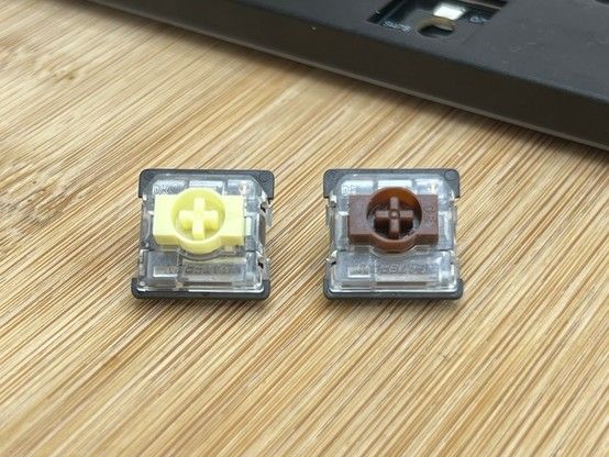 Two mechanical keyboard switches on a wooden surface, one with a yellow stem and the other with a brown stem, with part of a keyboard in the background.
