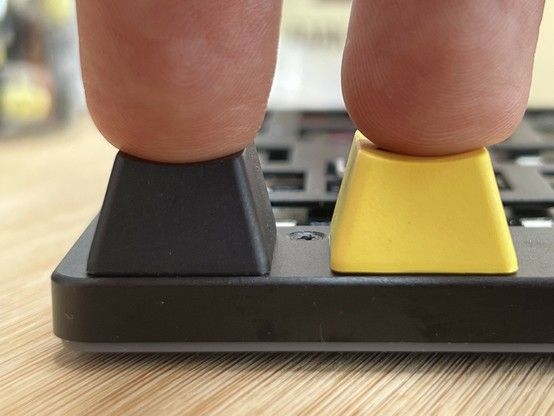 Close-up of two fingers pressing a black and a yellow key on a mechanical keyboard. Both are depressed fully and almost touching the surface of the keyboard.